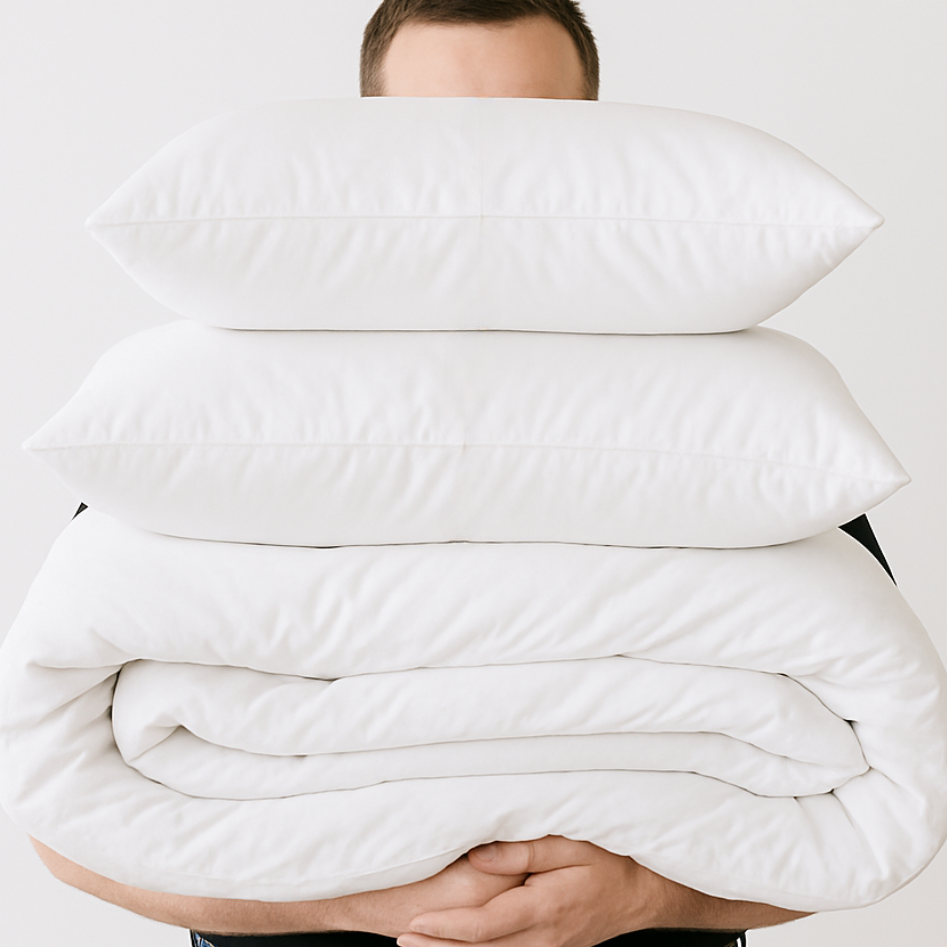 Person holding a stack of white pillows and quilt against a plain background