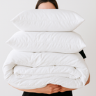 Person holding a stack of white pillows and a quilt against a plain background