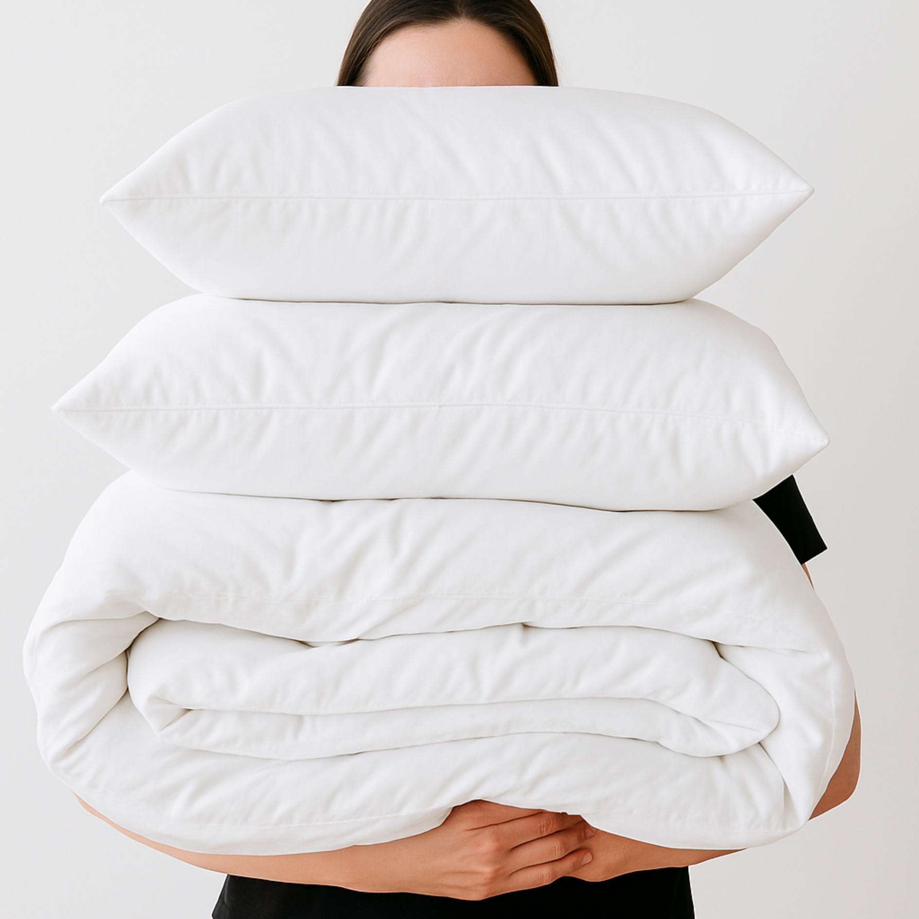 Person holding a stack of white pillows and a quilt against a plain background